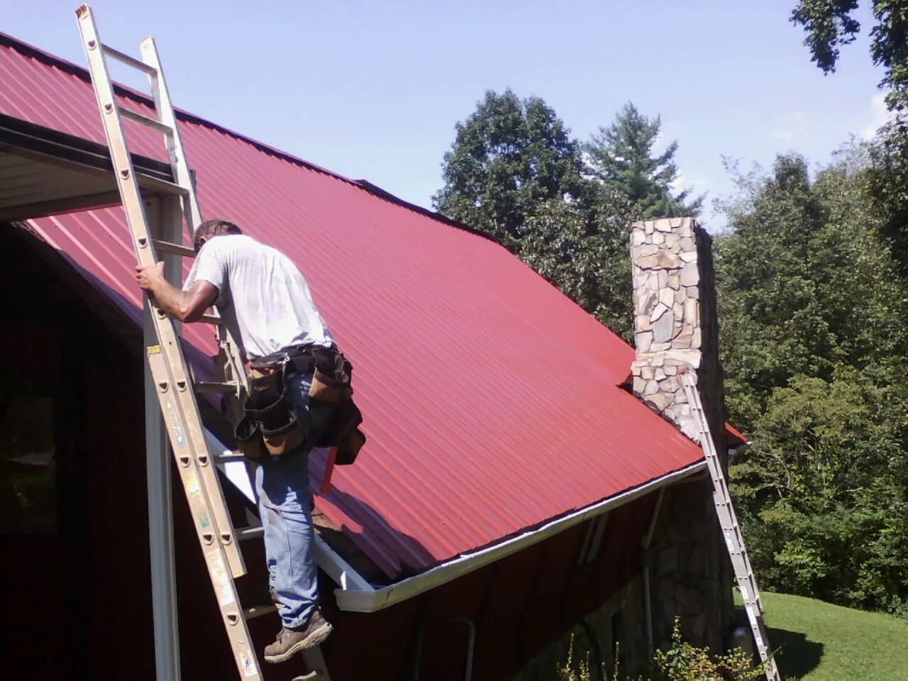Roofer on a ladder beside a red metal roof and stone chimney — Soffit & Fascia Repair by Roofing Brothers in Popejoy